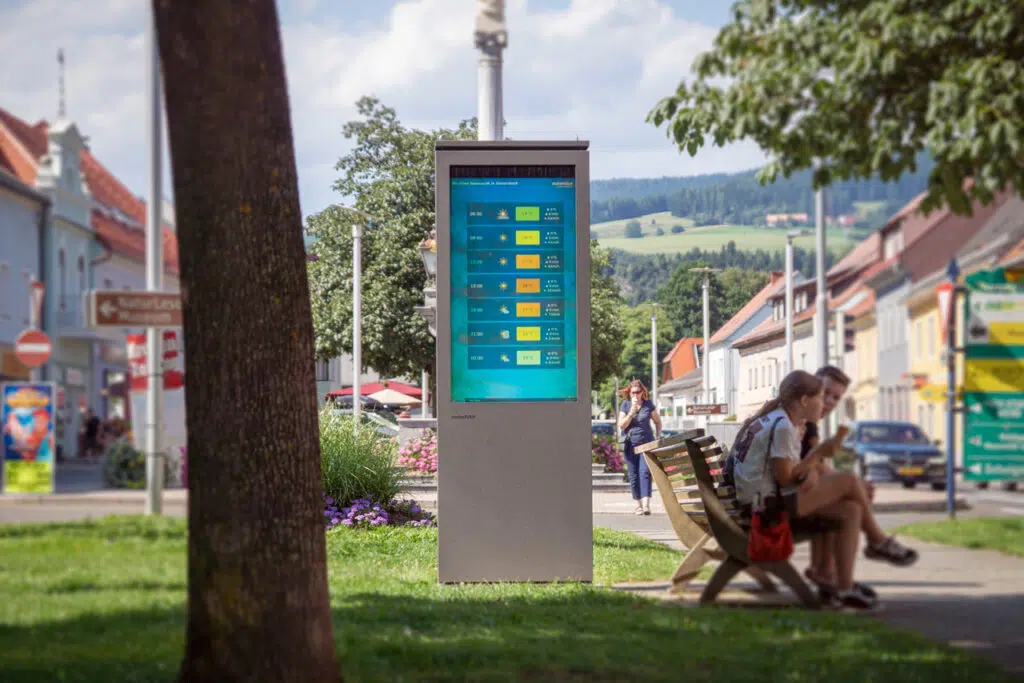Eine Outdoor Stele neben eine Parkbank auf der Personen sitzen und ein Baum steht links im Vordergrund