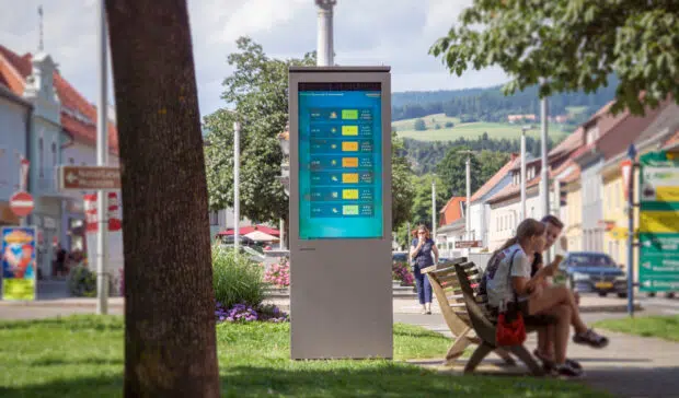 Eine Outdoor Stele neben eine Parkbank auf der Personen sitzen und ein Baum steht links im Vordergrund