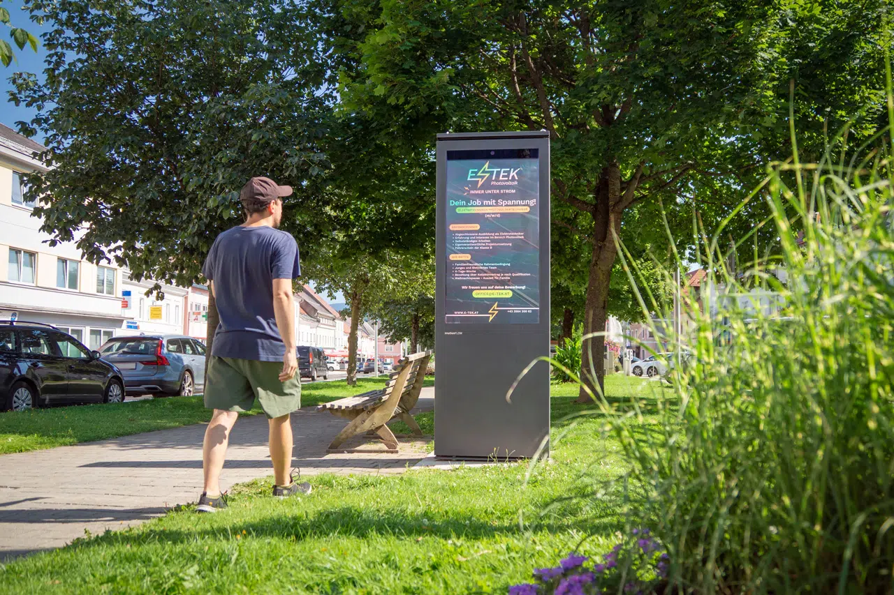 Eine Person steht neben einem Outdoor Monitor am Gehweg in einem Park