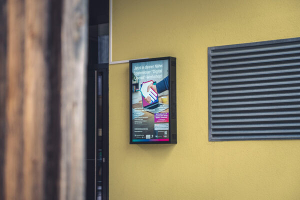 Ein Outdoor Screen hängend an einer gelben Wand vor einem Gebäude.