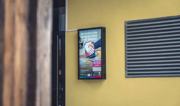 Ein Outdoor Screen hängend an einer gelben Wand vor einem Gebäude.