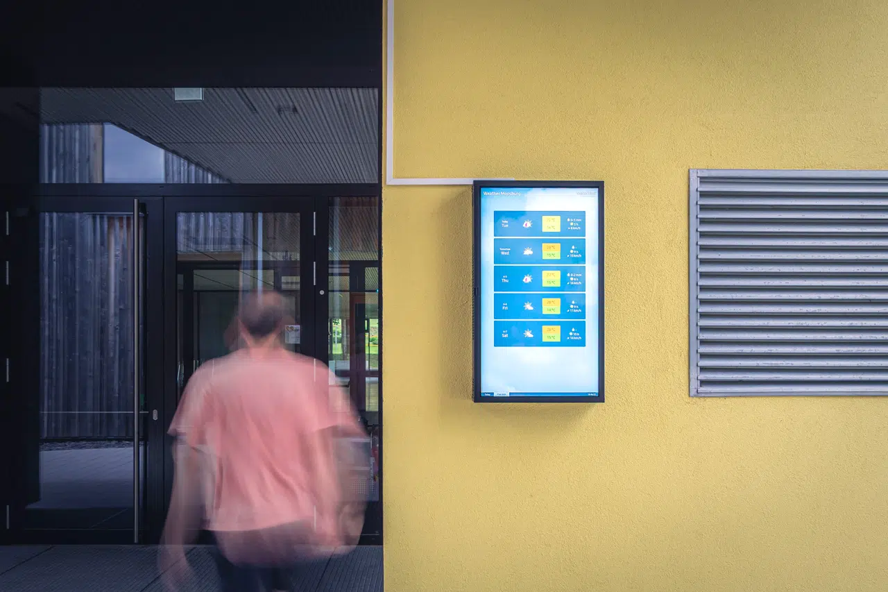 Ein Outdoor Screen hängend an einer gelben Wand vor einem Gebäude. Links eine Person bewegt sich auf den Eingang zu.