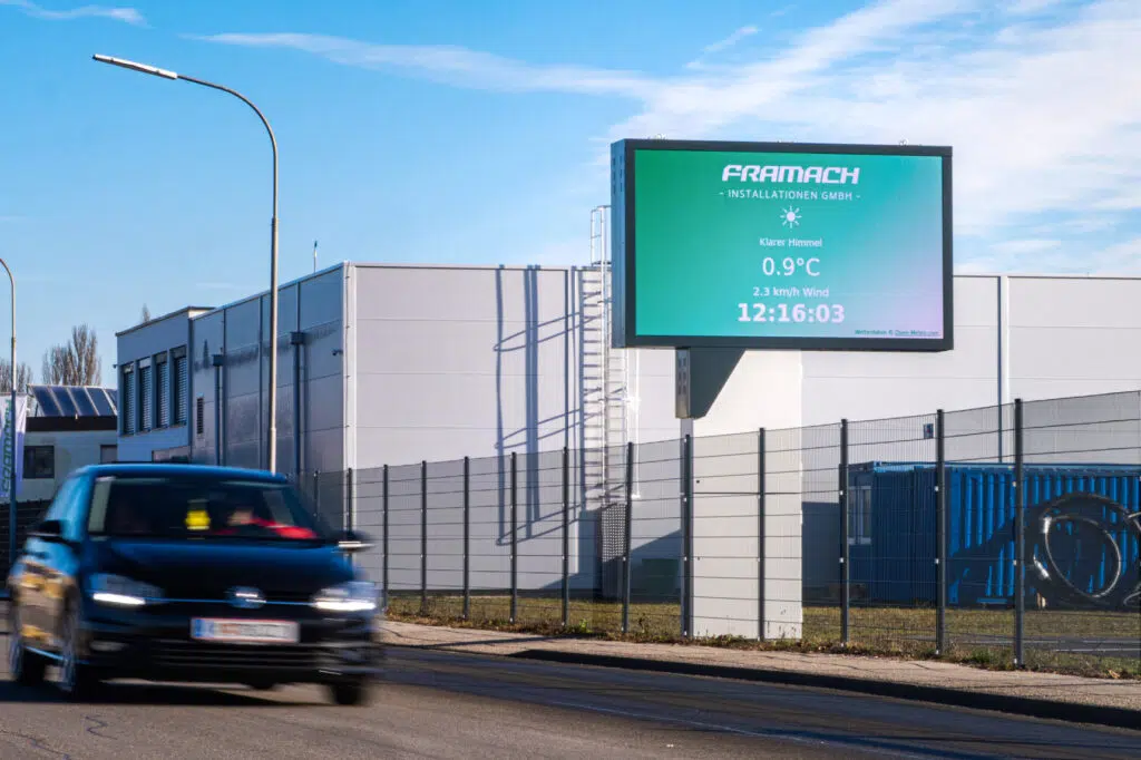freihstehende-led-wall-framach-wallerie Freistehende LED-Wall von Wallerie beim Unternehmen Framach Installationen GmbH in Framach, montiert auf einem Stahlmast neben dem Firmengebäude. Die Anzeige zeigt Wetter- und Uhrzeitinformationen. Im Vordergrund ein vorbeifahrendes Auto.