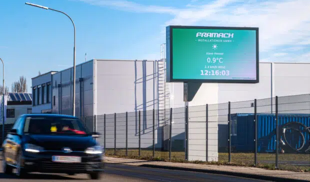 Freistehende LED-Wall von Wallerie beim Unternehmen Framach Installationen GmbH in Framach, montiert auf einem Stahlmast neben dem Firmengebäude. Die Anzeige zeigt Wetter- und Uhrzeitinformationen. Im Vordergrund ein vorbeifahrendes Auto.