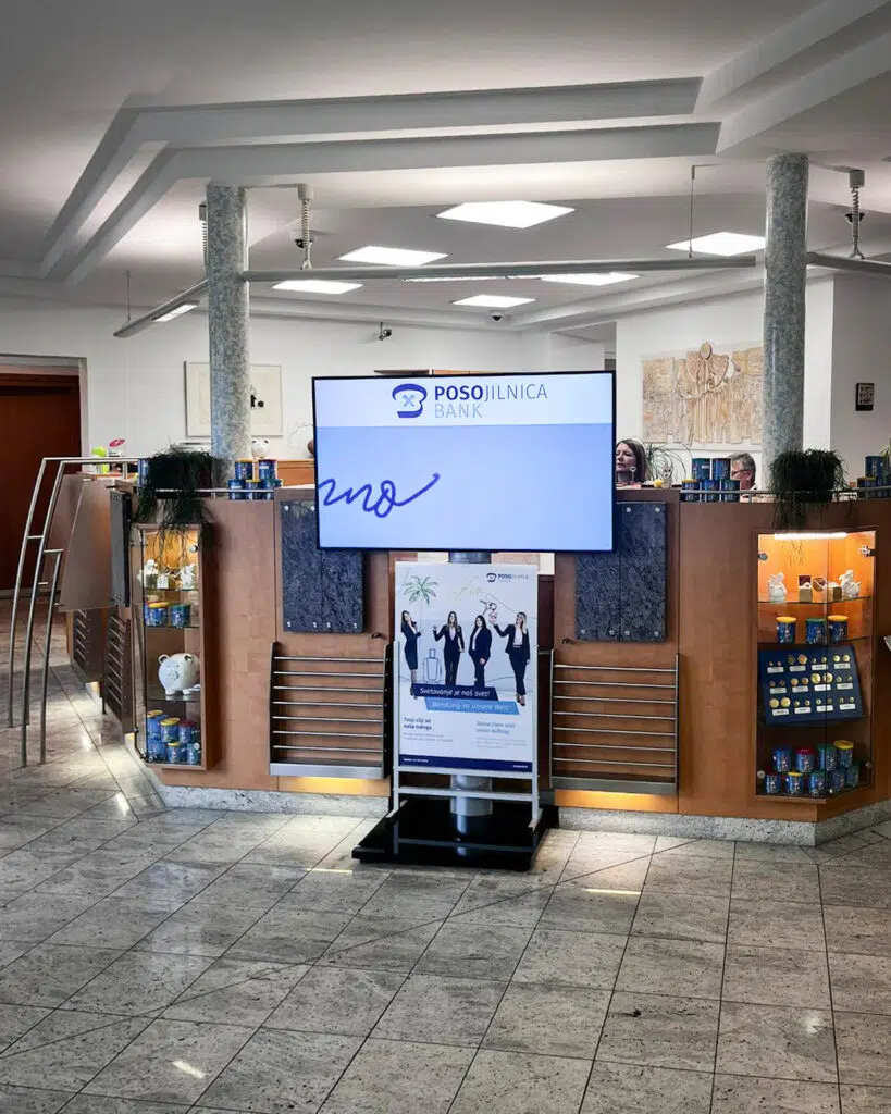 poso-bank-managed-digital-signage-system-by-wallerie-04 Digital Signage Installation im Eingangsbereich einer Posojilnica Bank-Filiale: Großer Wandscreen mit Posojilnica-Logo sowie ein freistehender Digital-Signage-Aufsteller mit Werbemotiv.